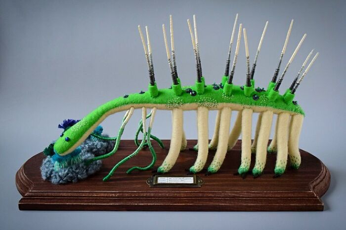 Felted artwork of a whimsical green creature with multiple legs and antennae displayed on a wooden base.