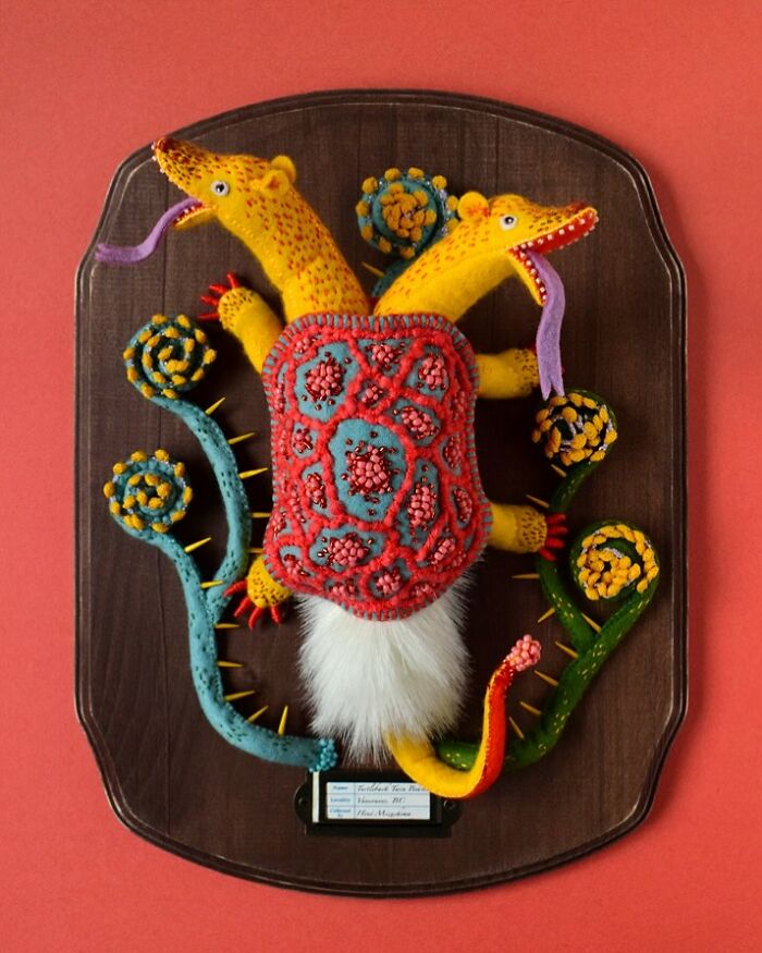 Felted artwork of a colorful two-headed creature mounted on a wooden plaque with whimsical felted plants.