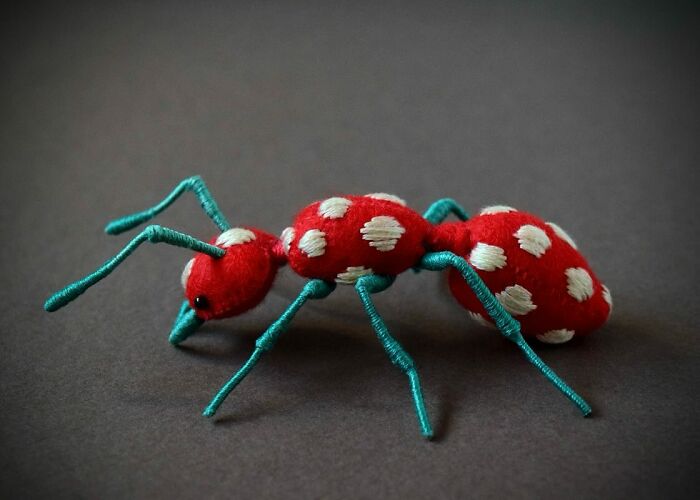 Red and white spotted felted ant artwork with blue wire legs displayed on a dark background, showcasing felted artworks detail.