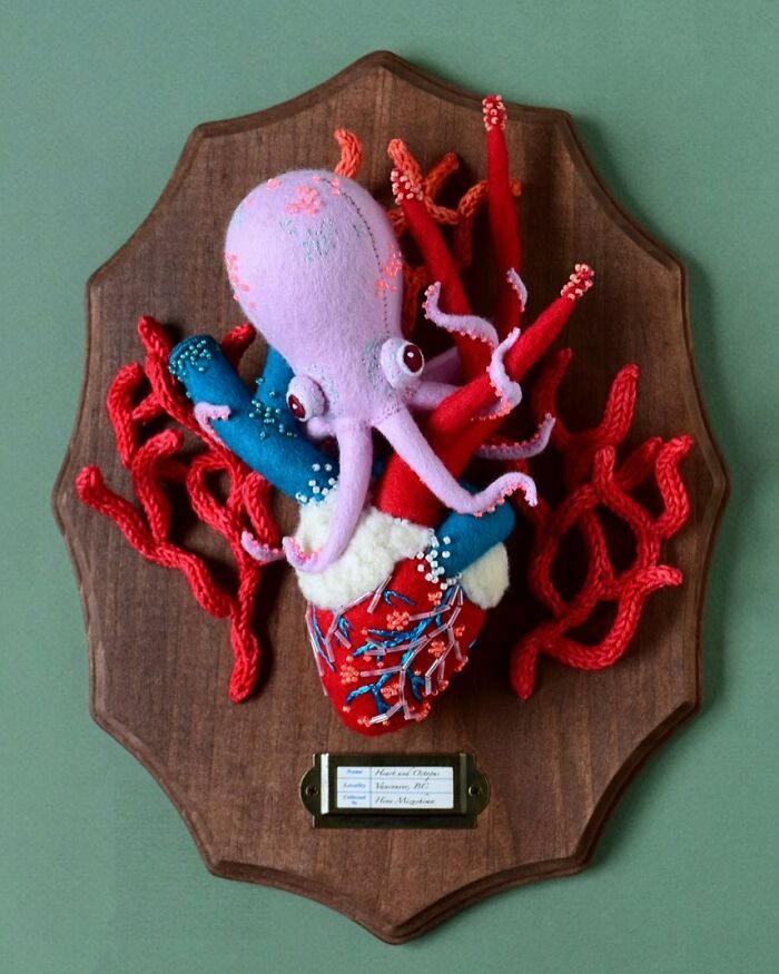Felted artwork of a pink octopus on a colorful heart with red coral, mounted on a wooden plaque against a green background.