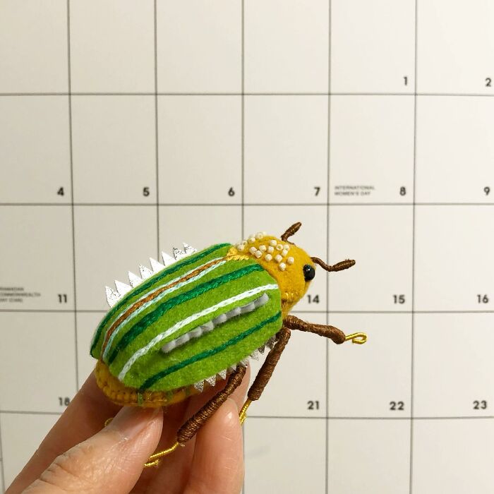 Hand holding a colorful felted beetle artwork with green and yellow stripes against a calendar background.