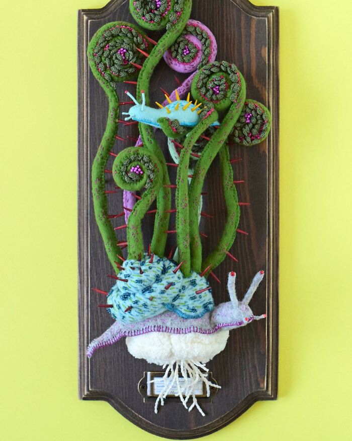 Felted artwork featuring a whimsical snail with vibrant green curly plants mounted on a wooden plaque.