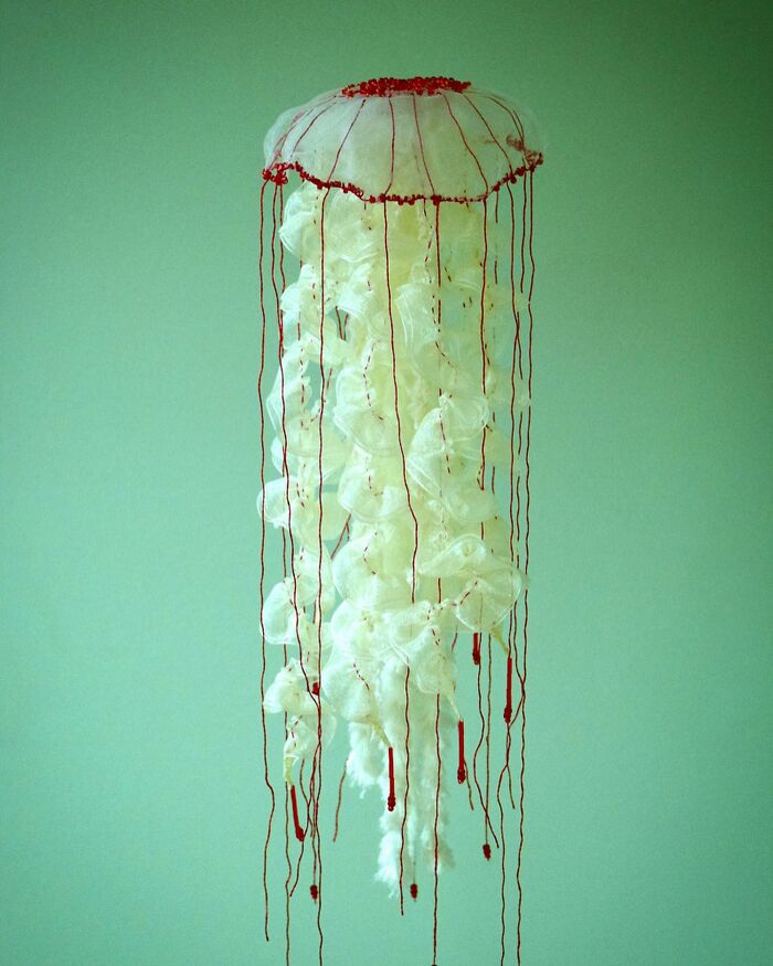 White and red felted artwork resembling a jellyfish with long thin red strands against a pale green background.
