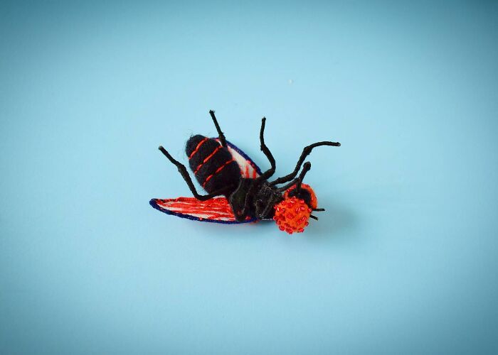 Felted artwork of a colorful insect with black, red, and blue details displayed against a light blue background.