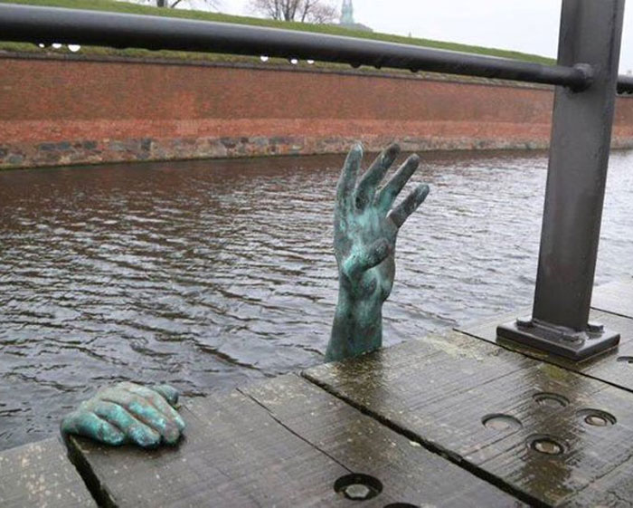Bronze sculpture of two hands emerging from water near a dock, representing a Danish prison cell symbol in public art.
