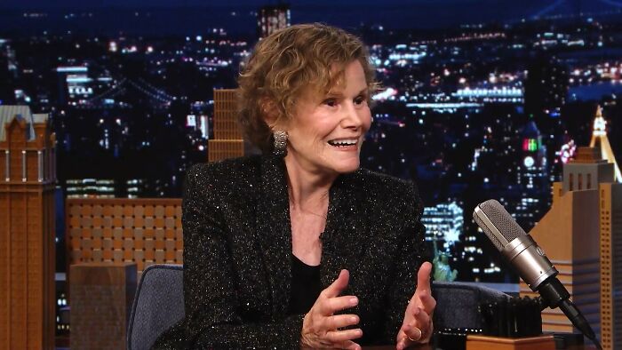 Woman sharing a joyful story about meeting an idol during a nighttime talk show interview with city lights in the background.