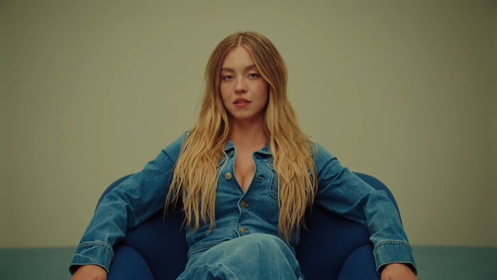 Young woman with long blonde hair wearing denim, sitting confidently on a chair, illustrating kaomojis cultural appropriation debate.