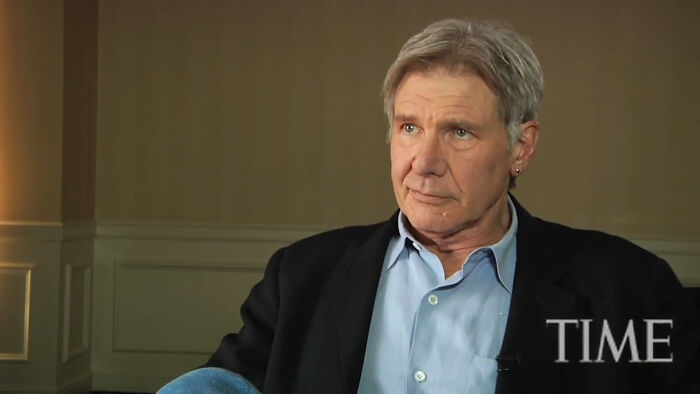 Man in a blue shirt and black jacket sharing an interview about meeting an idol, in a neutral indoor setting.
