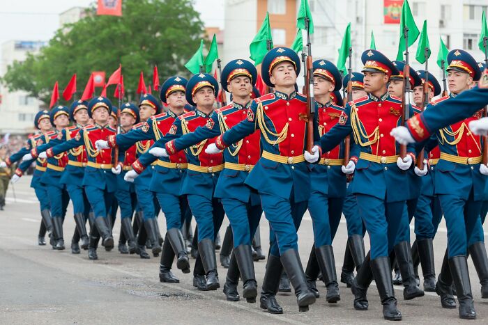 A group of soldiers marching in colorful uniforms and caps, holding flags during a parade in an urban setting.