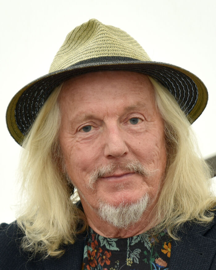Older man with long blond hair and a beard, wearing a beige hat and floral shirt, representing famous fraud cases.