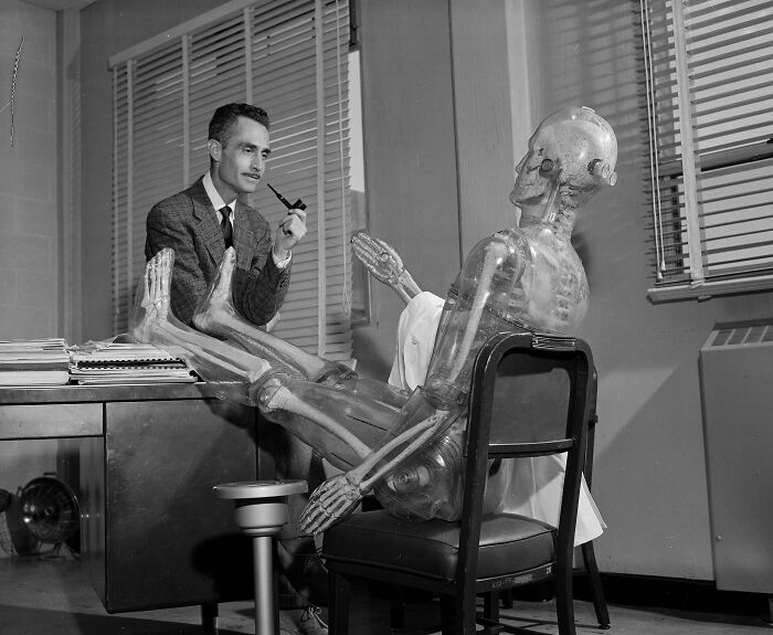 Man in suit smoking pipe while interacting with transparent medical skeleton model in old-school medical setting