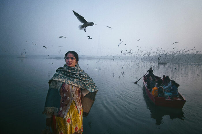 Woman wrapped in shawl standing near foggy waters with boat and birds, capturing candid moments of everyday life in Istanbul.