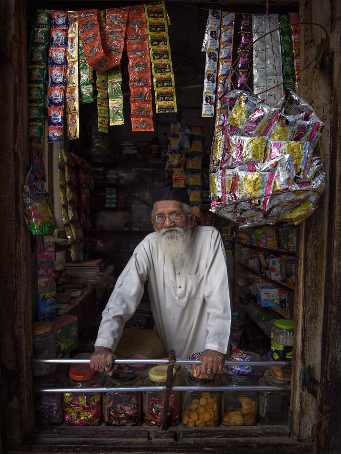 Elderly shopkeeper with white beard stands inside small store filled with colorful packets in candid moments of everyday life Istanbul.