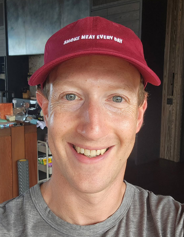 Mark Zuckerberg wearing red cap and gray shirt, smiling in a casual indoor setting related to hot mic moment.