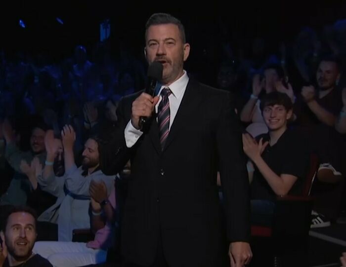 Jimmy Kimmel in a suit holding a microphone on stage with an audience applauding during a show recording. Jimmy Kimmel in a suit holding a microphone on stage with an audience applauding during a show recording.