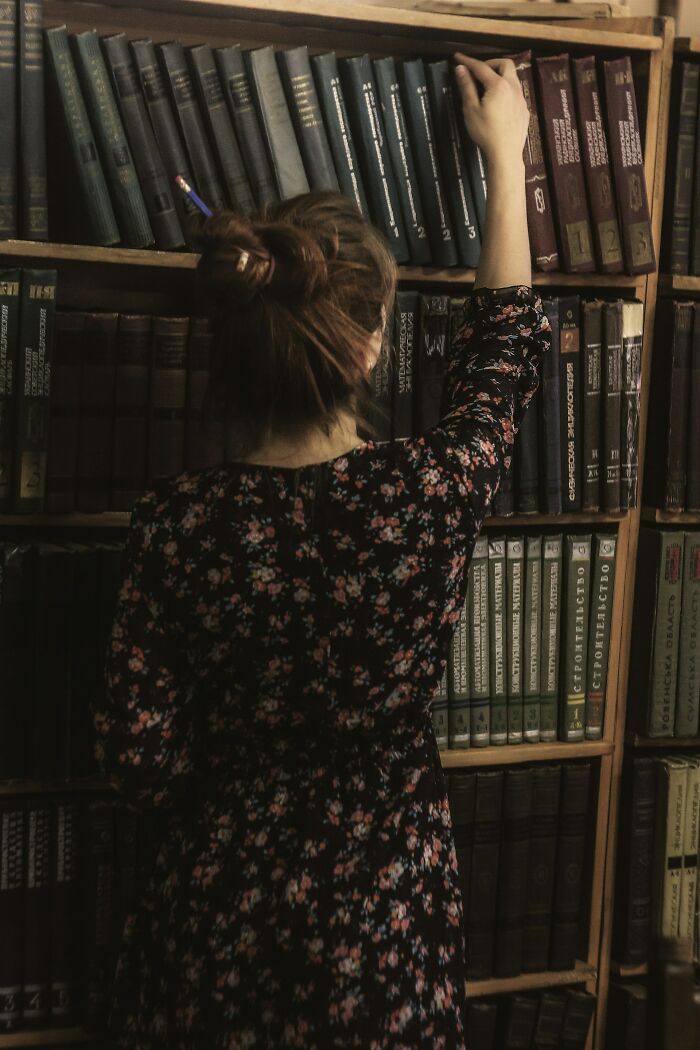 Woman in floral dress reaching for a book on a shelf in a dimly lit library representing jobs for ultra-wealthy people.
