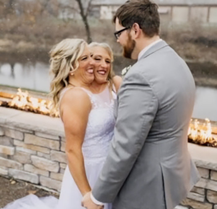 Conjoined twins Abby and Brittany in wedding attire holding hands with a man near a stone wall by a river.