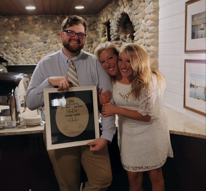 Three people smiling indoors, one holding a framed item, related to conjoined twins Abby and Brittany with a baby.