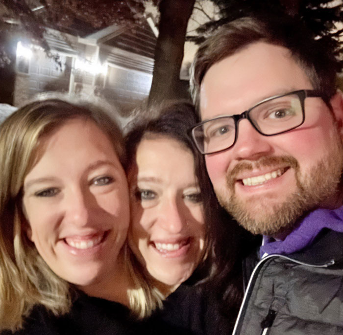 Conjoined twins Abby and Brittany smiling outdoors at night with a man, sparking expert insights on who would be the mom.