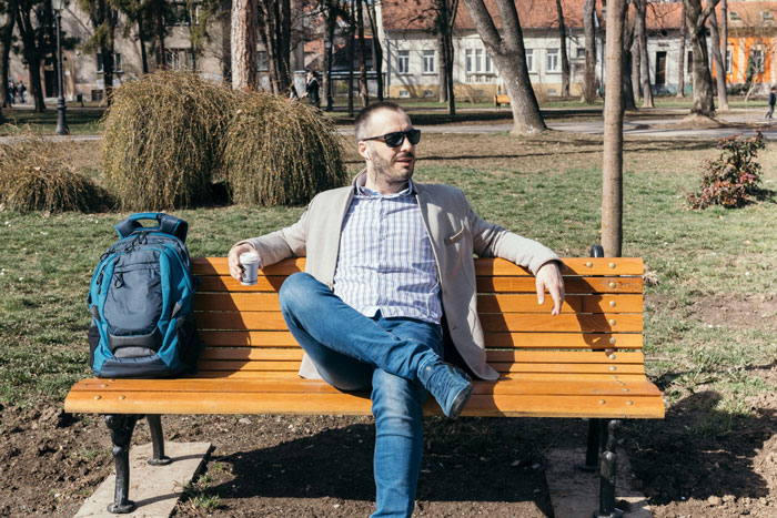Man wearing sunglasses sitting on a park bench with backpack nearby, in a no-walking area setting.