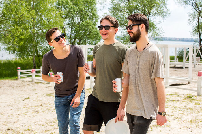 Three former high school stars wearing sunglasses, laughing and holding drinks outdoors by a lake on a sunny day.