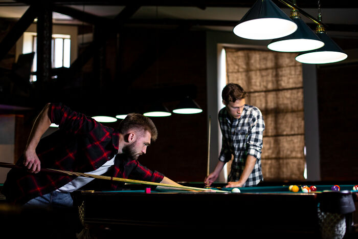 Two young men in casual shirts playing pool indoors, reflecting on where life took them after high school glory days.