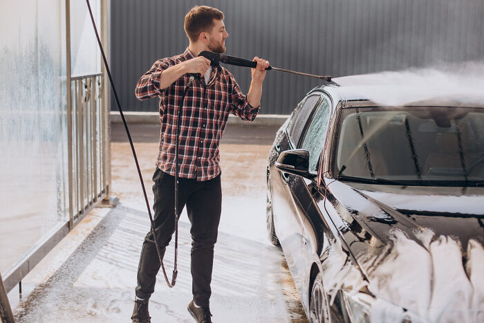Young man washing a black car with a pressure washer, illustrating life after the glory days of high school stars.