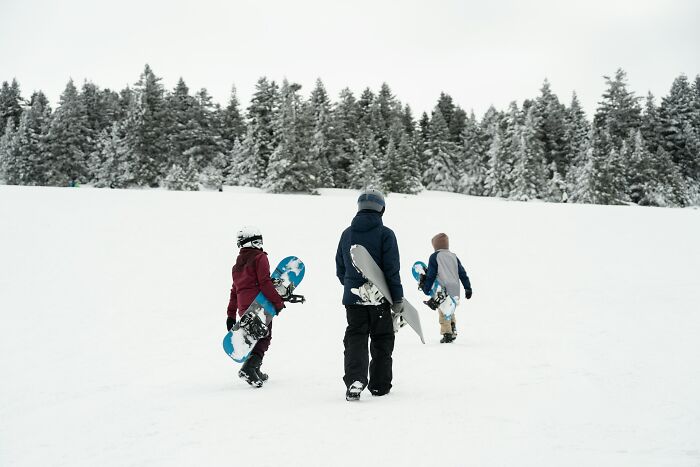 Three people carrying snowboards in a snowy landscape with trees, contrasting with complaints about traveling to the Bahamas.