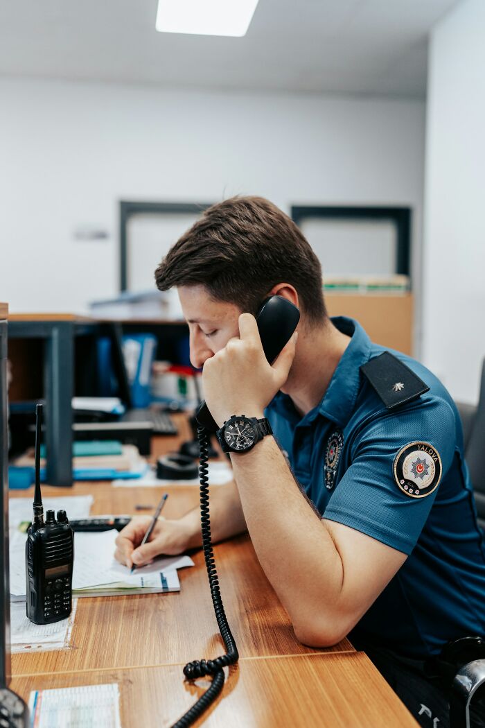 Police officer in uniform speaking on a landline phone and taking notes at a wooden desk in an office setting.