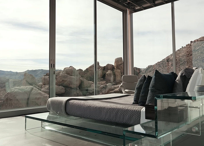 Modern bedroom interior with glass walls featuring rocky desert landscape in the famous invisible house Airbnb owners charge.