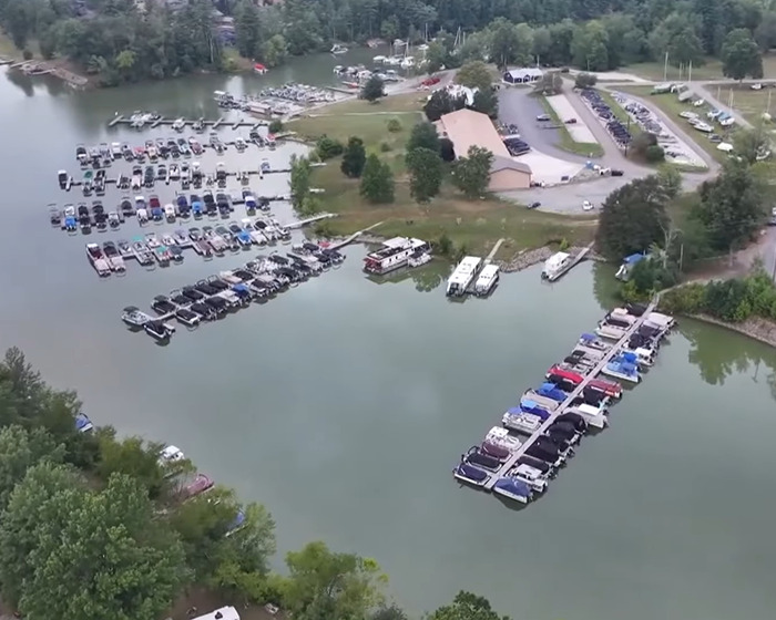 Aerial view of a boat marina surrounded by trees with vehicles parked near docks on a calm lake.
