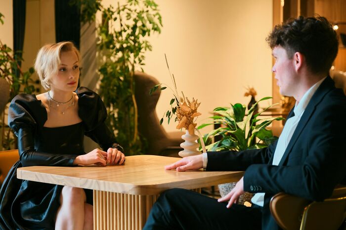 Two people in formal attire having a tense conversation at a restaurant, servers overhearing crazy relationship stories.