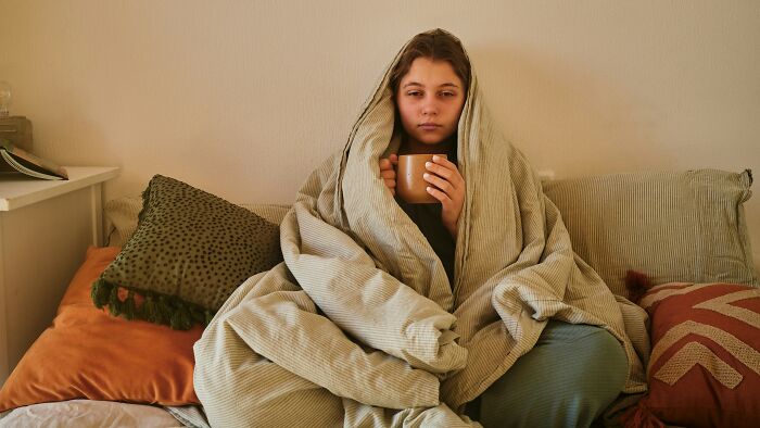 Sick woman wrapped in blanket, holding a mug and looking unsure about attending wedding with positive Covid test.