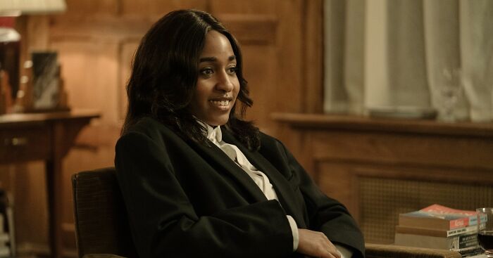 Ayo Edebiri seated comfortably in a dark blazer, smiling during a casual indoor interview setting with wooden background.