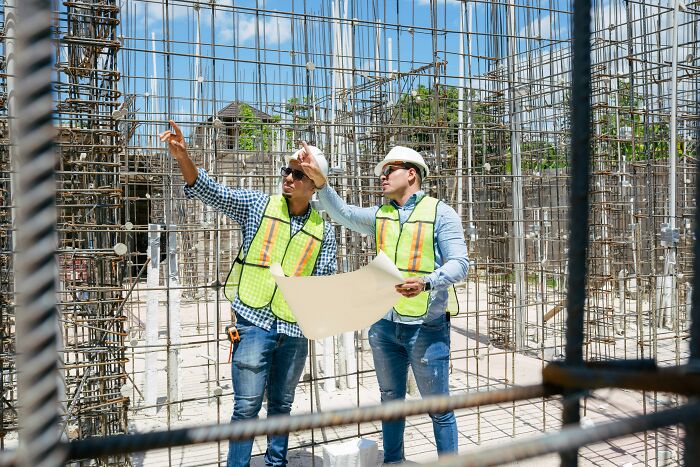 Two construction workers wearing safety vests and helmets reviewing building plans inside metal framework, professions not relationship material.