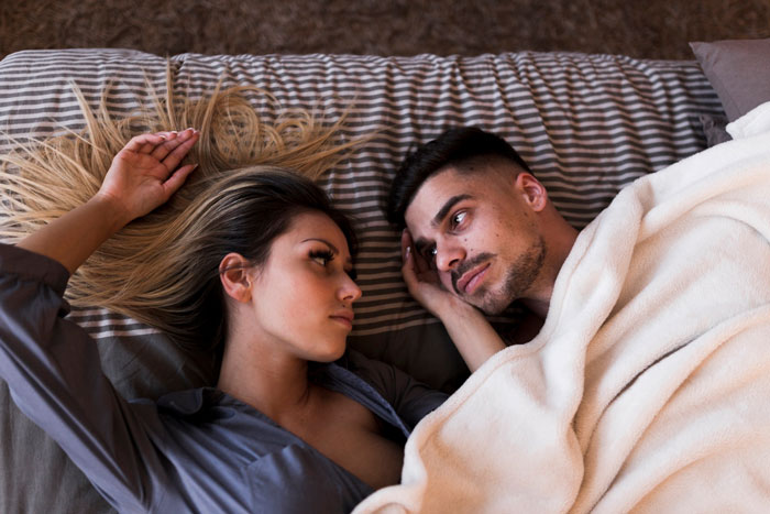 Couple lying in bed together, the man looking at his wife, referencing throuple and anime pillow concerns. Couple lying in bed together, the man looking at his wife, referencing throuple and anime pillow concerns.