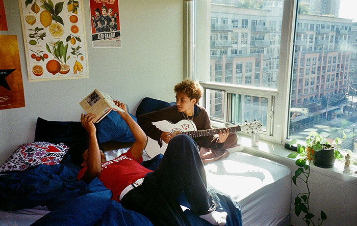 Two young men in a bright room, one playing guitar and the other reading, highlighting awkward boxer moments.