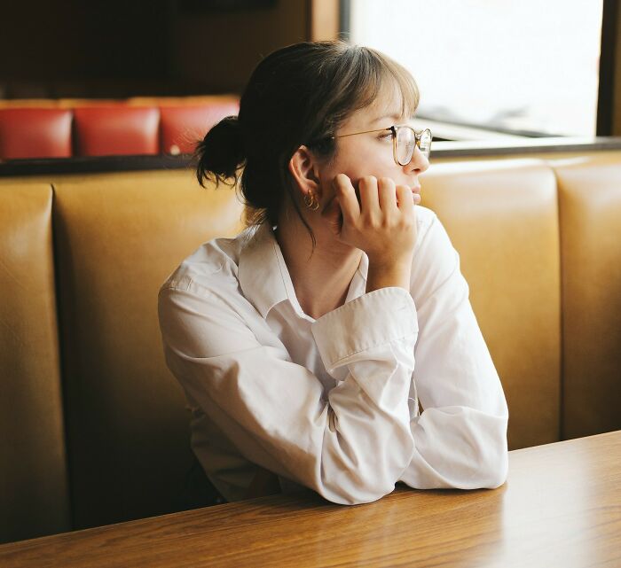 Young woman with glasses sitting in a booth, looking away thoughtfully, reflecting on crazy stories servers overheard.