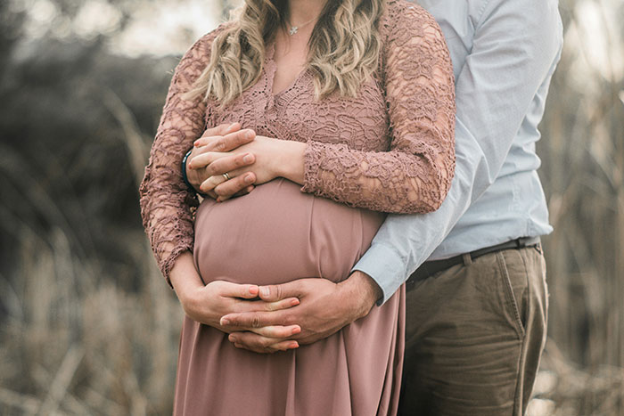 Pregnant woman in a pink lace dress embraced by husband outdoors, highlighting love and complex relationship emotions.