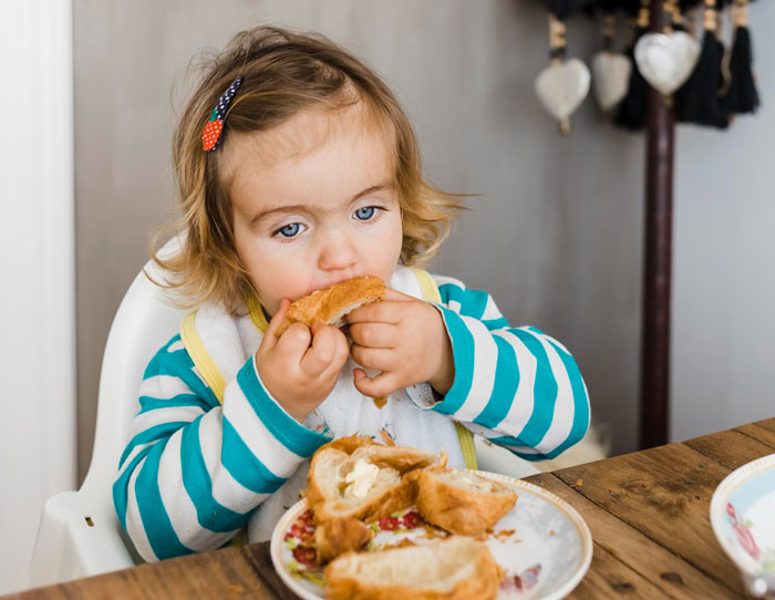 Toddler girl eating bread at a wooden table, highlighting concerns about eating disorder comments from ex and dad's reaction.