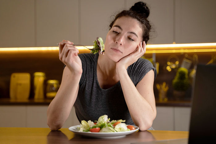 Woman in kitchen looking disinterested at salad, illustrating eating disorder behavior and emotional struggle with food.