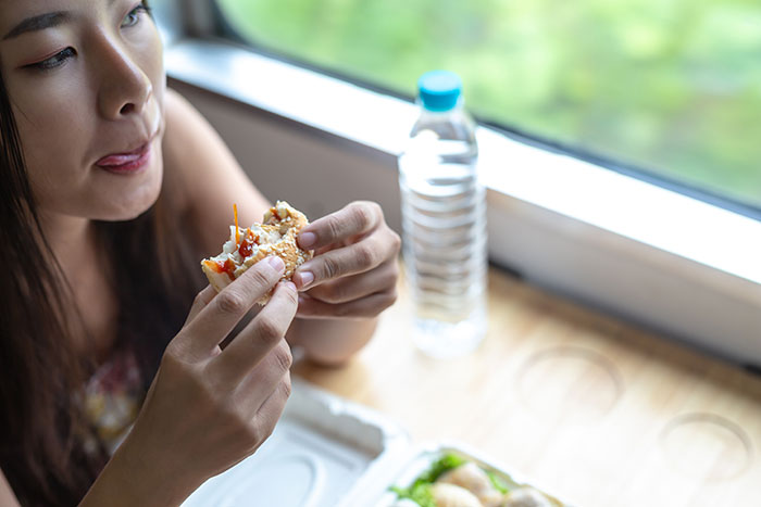 Woman eating a sandwich near a window, highlighting risks of ignoring child&rsquo;s severe allergy leading to emergency visit.
