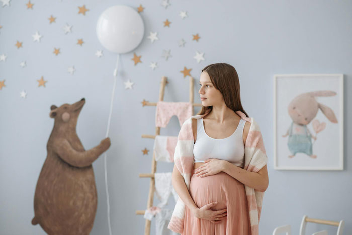 Pregnant woman in nursery holding belly, looking thoughtful with baby room decor including bear and bunny artwork behind her.