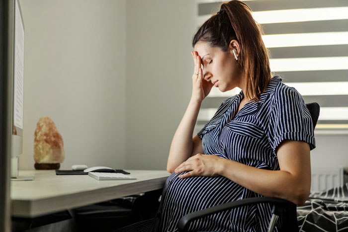 Pregnant woman distressed sitting at a desk, holding her belly and forehead, shocked by hubby&rsquo;s affair and matching baby name.