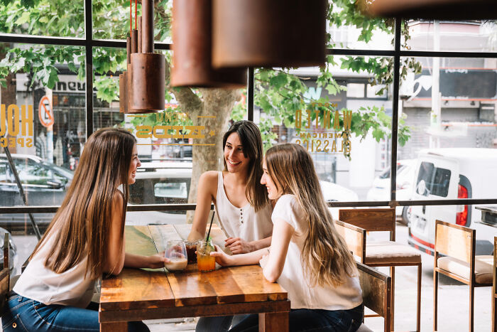 Three women sitting at a wooden table in a cafe, chatting and enjoying drinks in a bright, leafy setting.