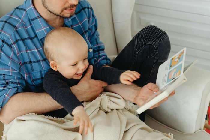 Dad taking care of son for the first time, reading a book while holding baby wrapped in a beige blanket at home.