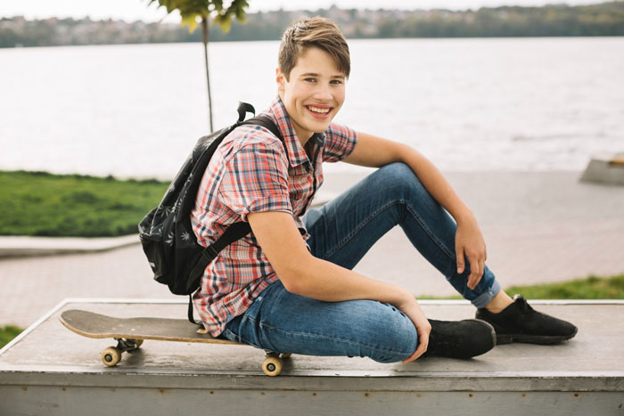 Teen boy with backpack sitting on skateboard by the water, representing woman&rsquo;s teen bro who is a nightmare to deal with.