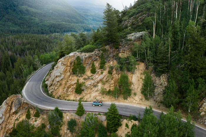 A winding mountain road with a vehicle driving along sharp curves surrounded by dense forest landscape and rocky cliffs.