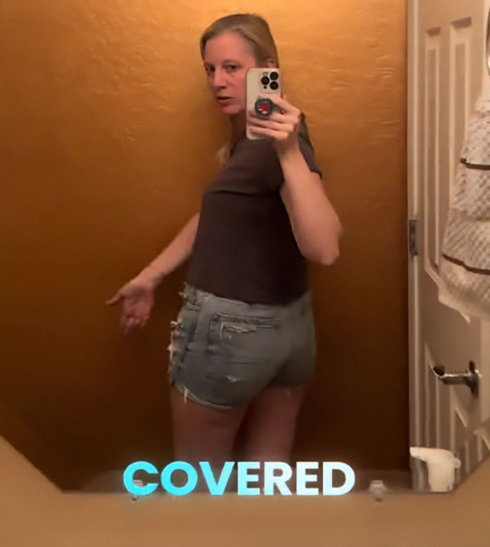 Woman taking mirror selfie wearing distressed mom shorts and dark top in a bathroom with brown walls.