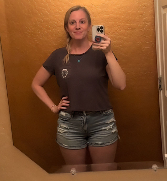 Woman taking a mirror selfie wearing distressed mom shorts and a casual dark t-shirt in a textured bathroom.
